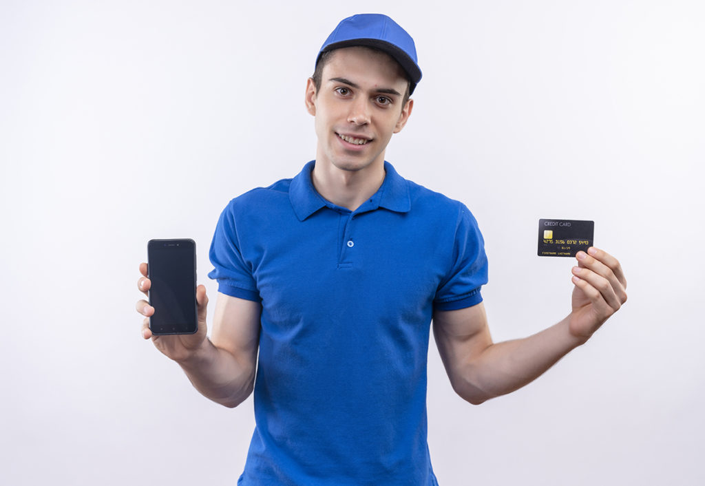 young courier wearing blue uniform and blue cap shows phone and card over white isolated background